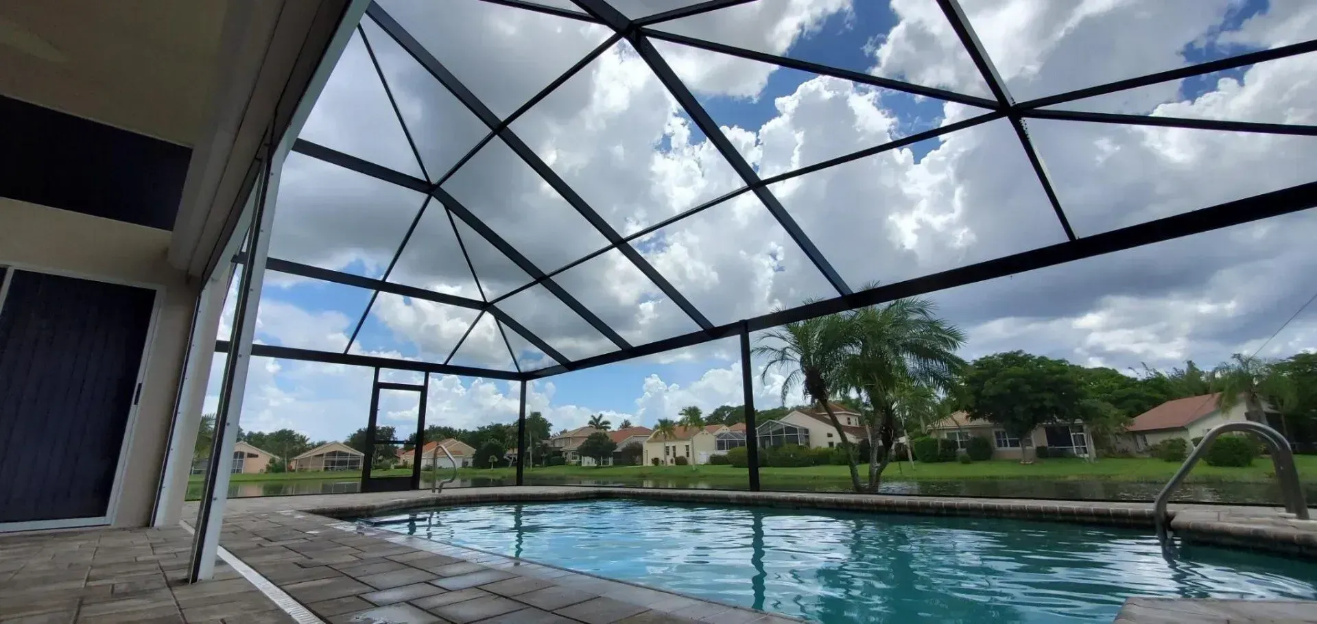 Pool under a screened enclosure with houses in the background and a cloudy sky.