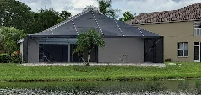 A grey screened-in patio, attached to a light-colored house, sits next to a pond.