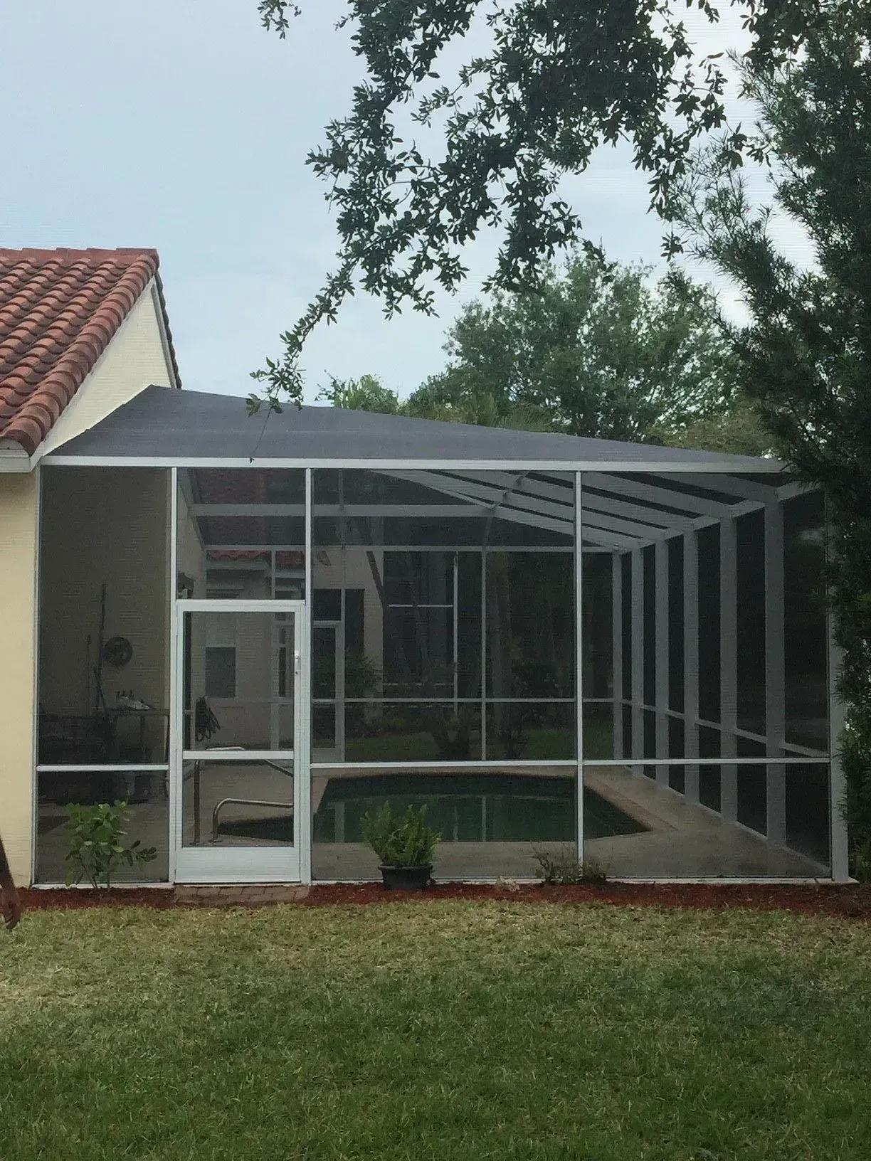 Screened-in pool enclosure with a door, white frame, black roof, and adjacent to a house with an orange tile roof.