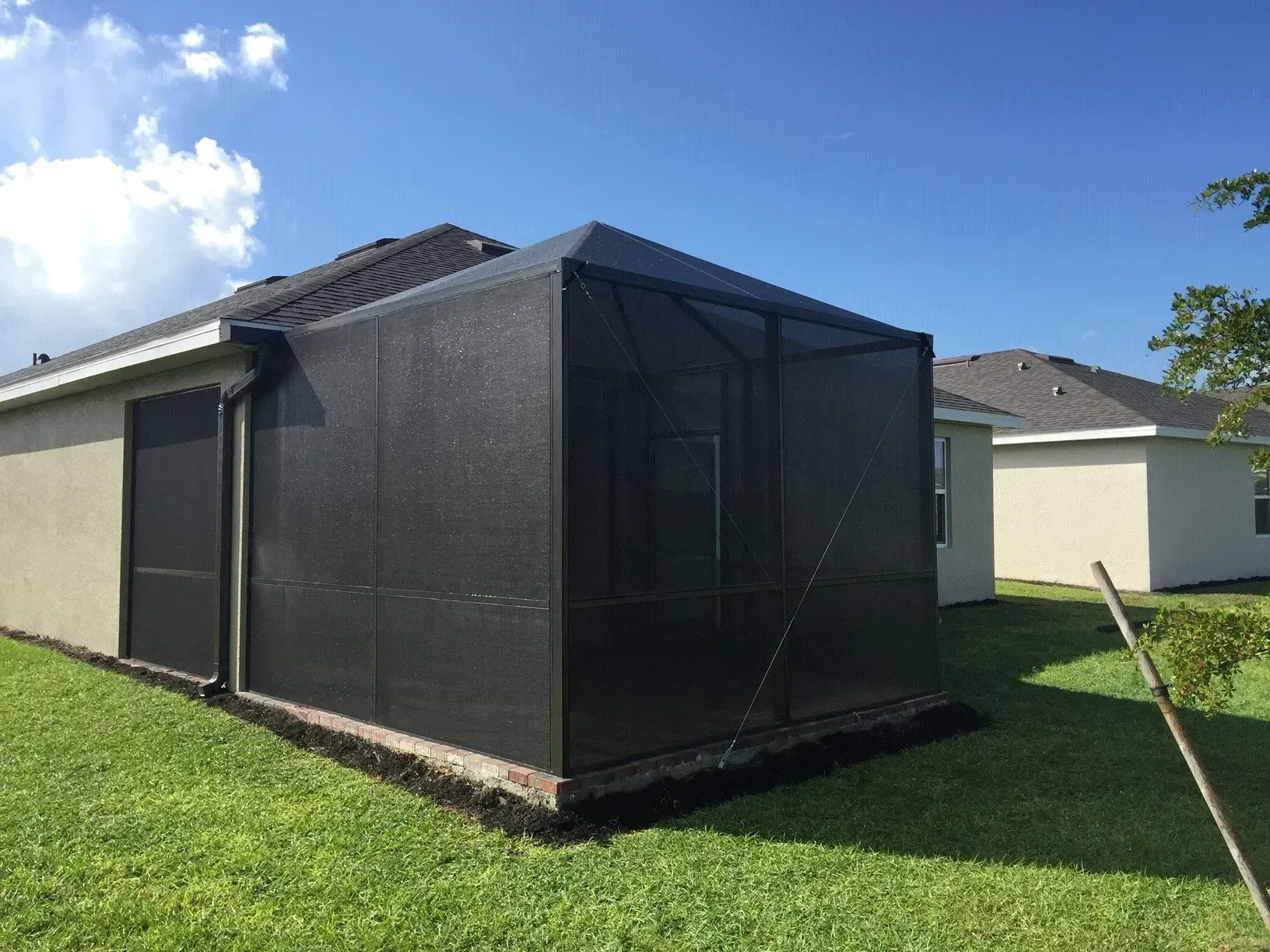 Screened-in porch attached to a beige house, with black screens and roof, on green grass under a blue sky.