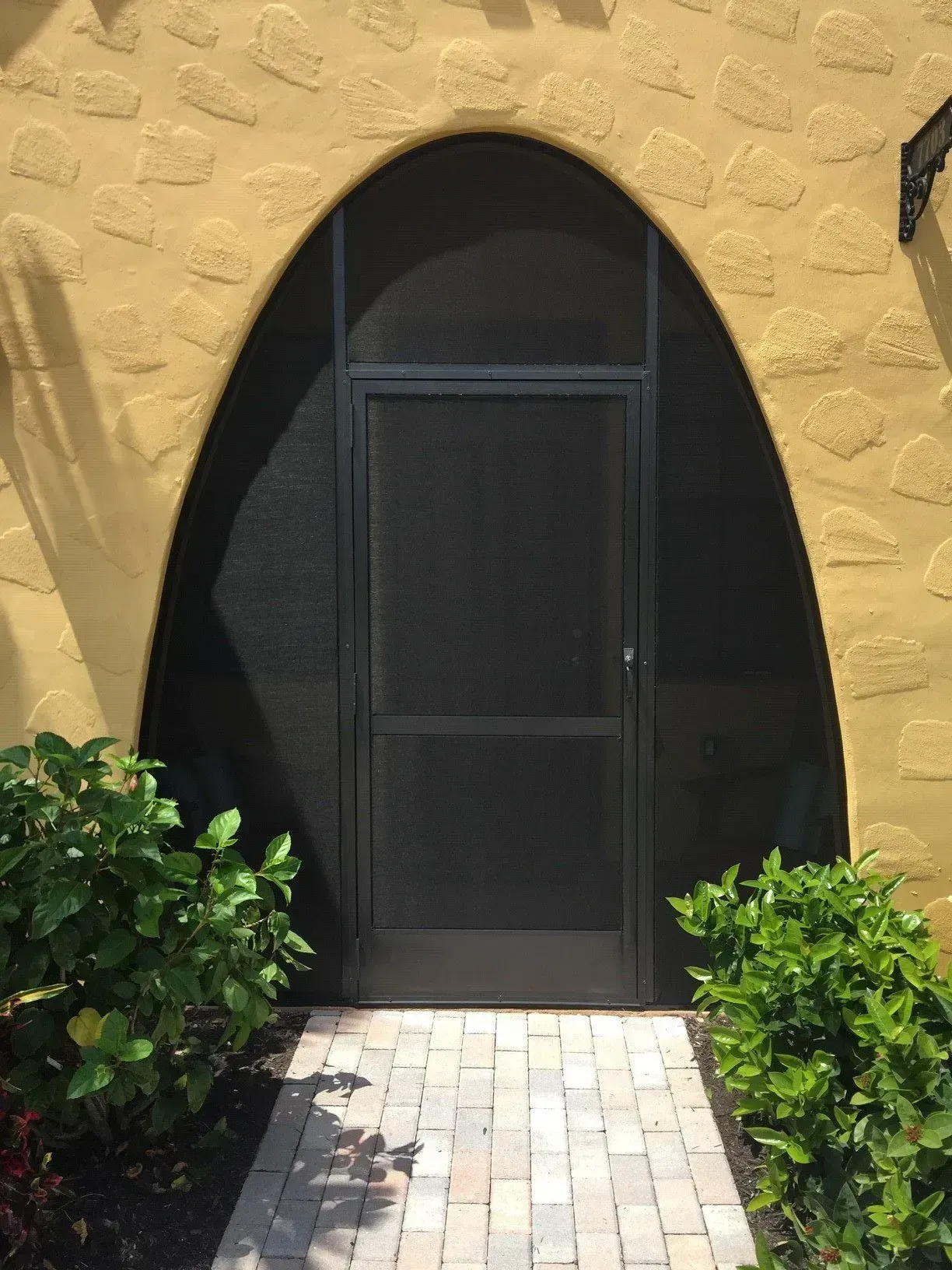 Black screen door in arched entryway of a building with textured, yellow stucco and a brick pathway.