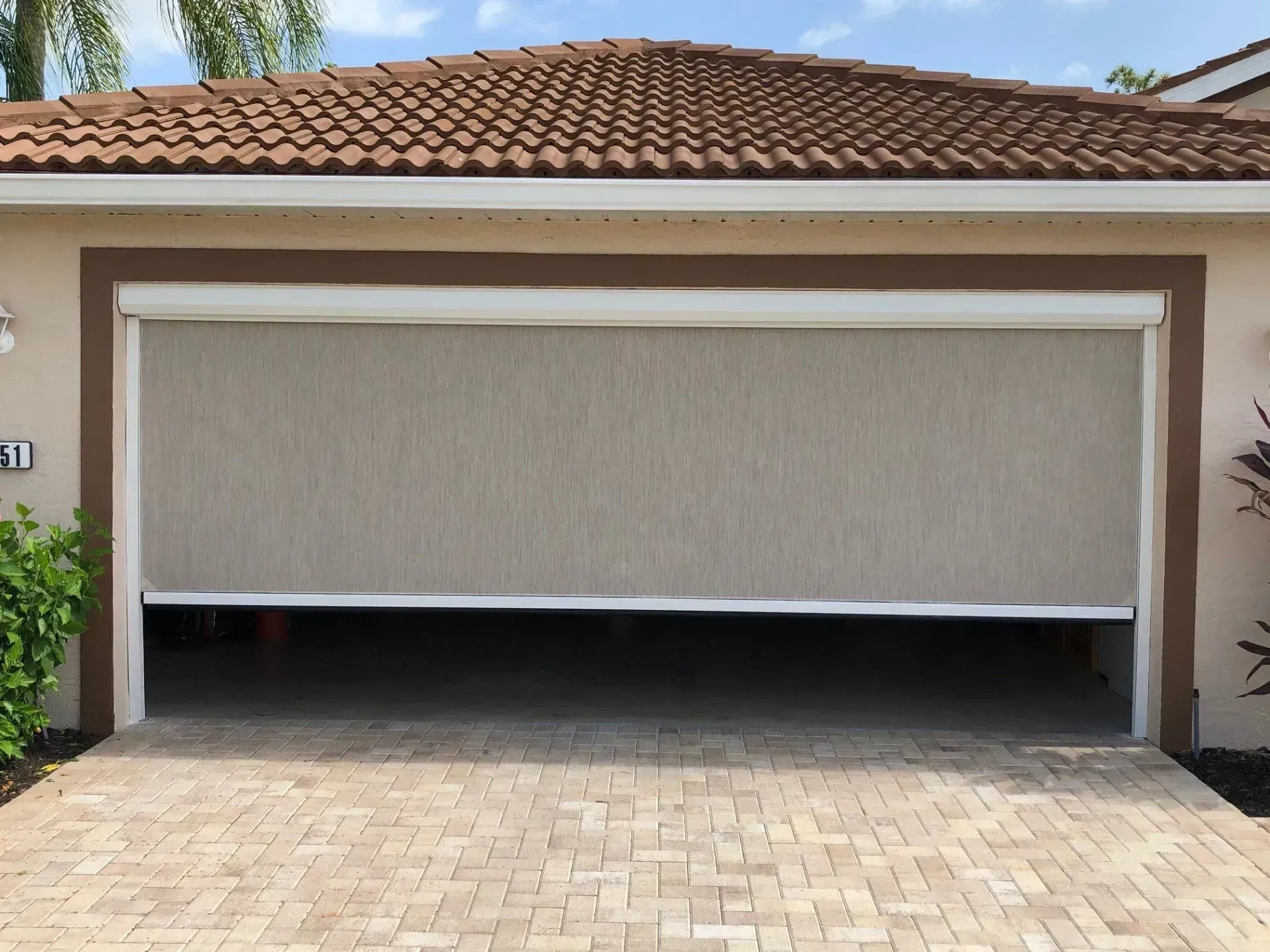 Garage with beige roll-down screen partially lowered; tan brick driveway, brown trim, and terracotta roof.