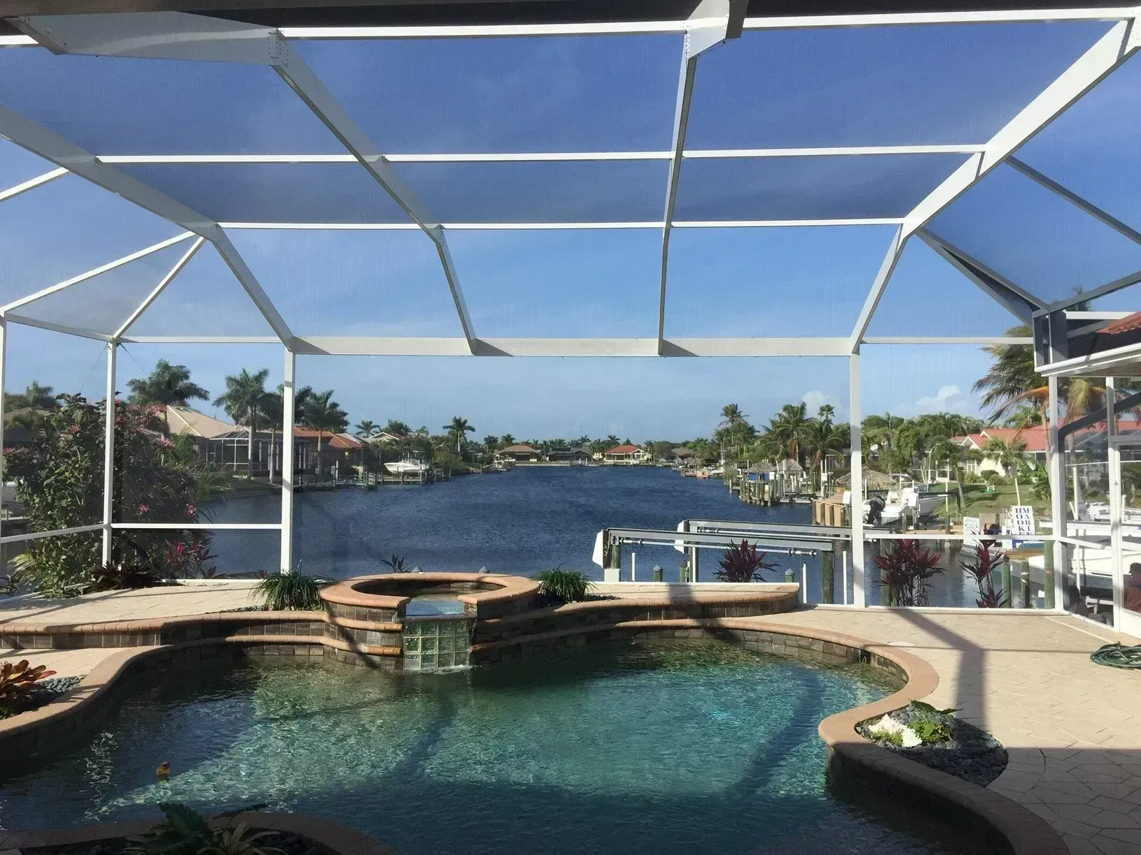 Pool with waterfall and screened enclosure overlooking a canal with houses and boats, under a blue sky.