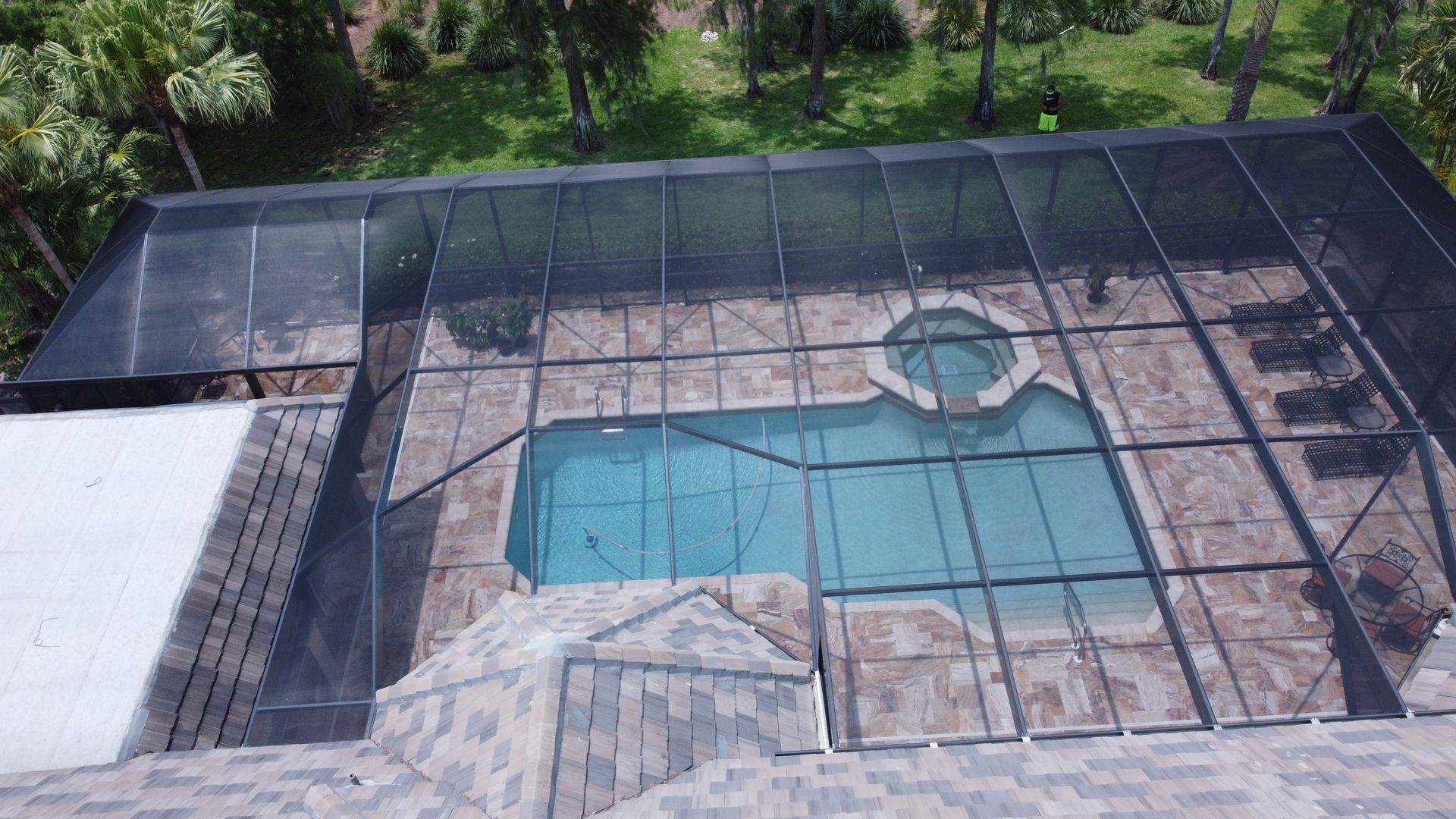 Overhead view of a swimming pool with screen enclosure on a stone patio, with surrounding green trees.