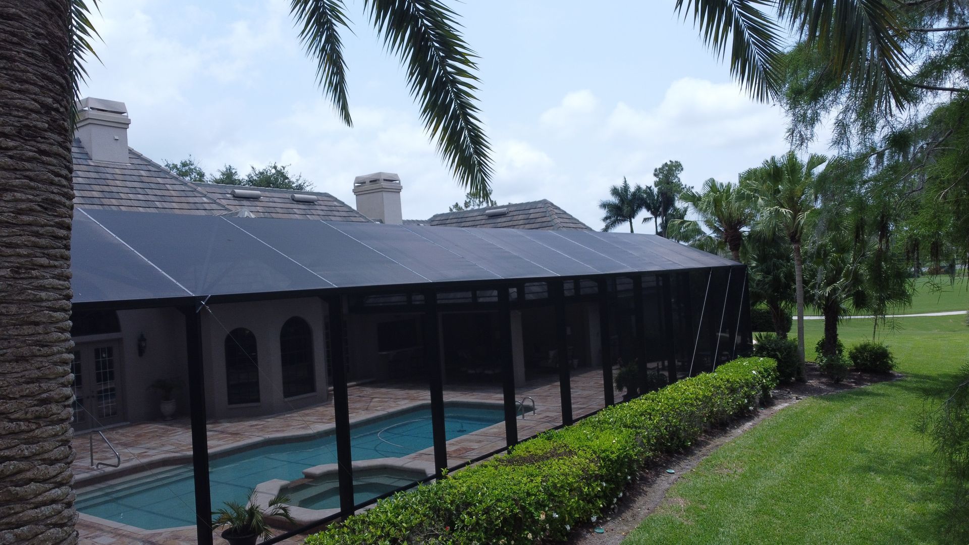 Pool area with solar panels on screen enclosure roof; green lawn, palm trees, blue sky.