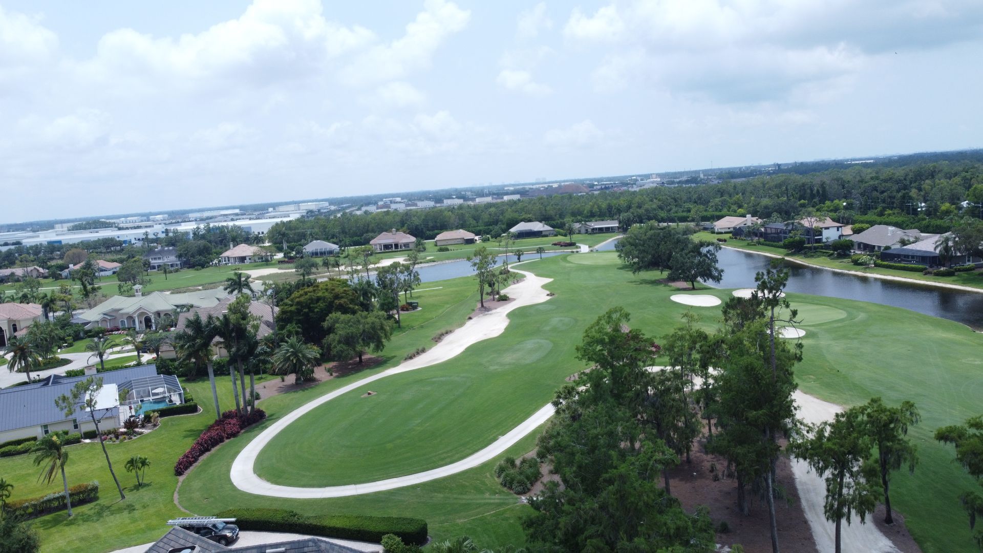 Aerial view of a green golf course with a few houses and a lake. Cloudy sky overhead.
