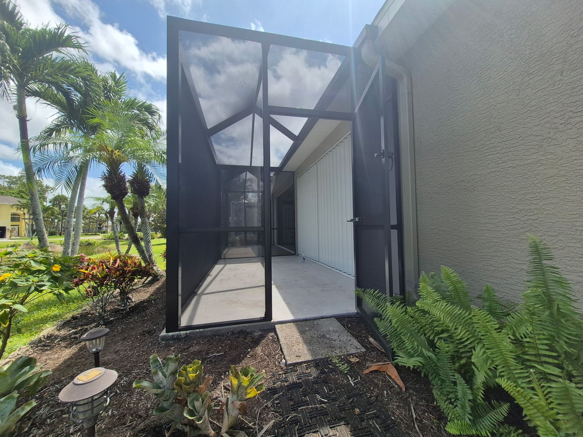 Screened-in patio with open doors, beside a light-colored building. Tropical plants and palm trees are visible.