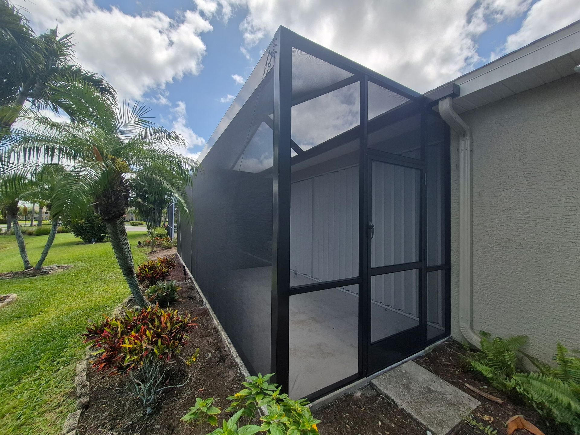 Screened-in porch attached to a light-colored house. Black frame with dark screens, surrounded by plants and grass.