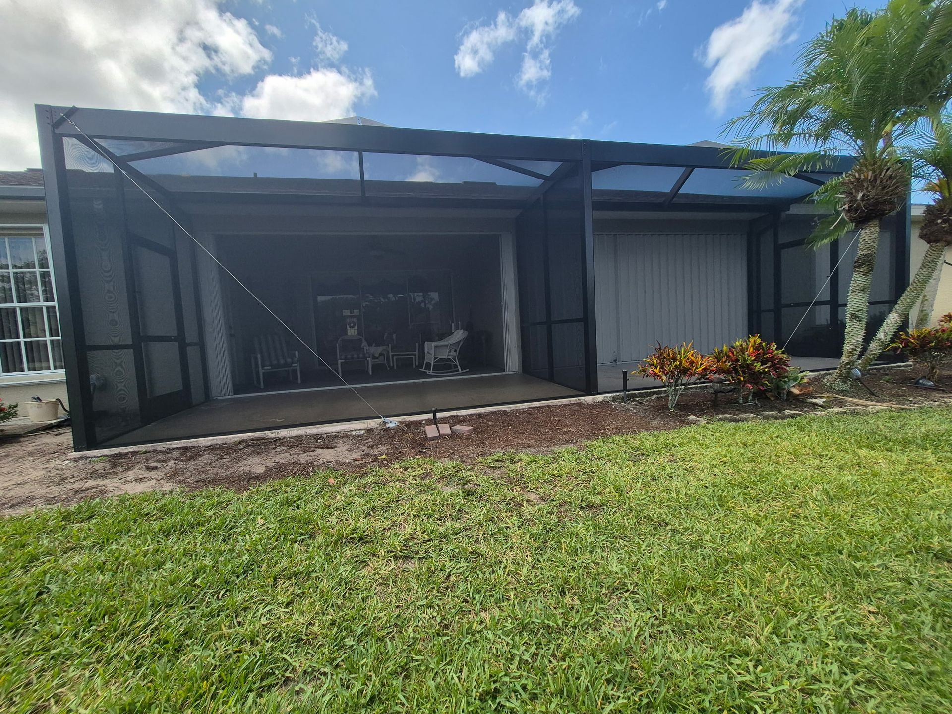 Screened-in patio with dark framing and screens, attached to a light-colored building, with green grass in the foreground.