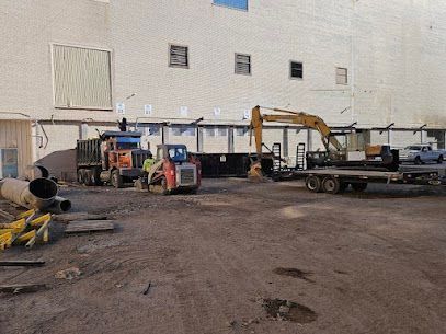 Construction yard with excavators, trucks, and equipment beside a large white building.