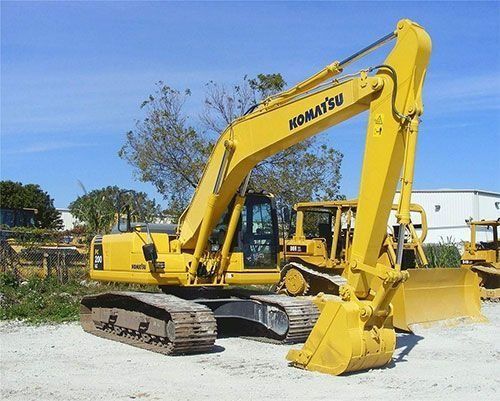 Yellow Komatsu excavator parked on gravel near trees and buildings under a blue sky
