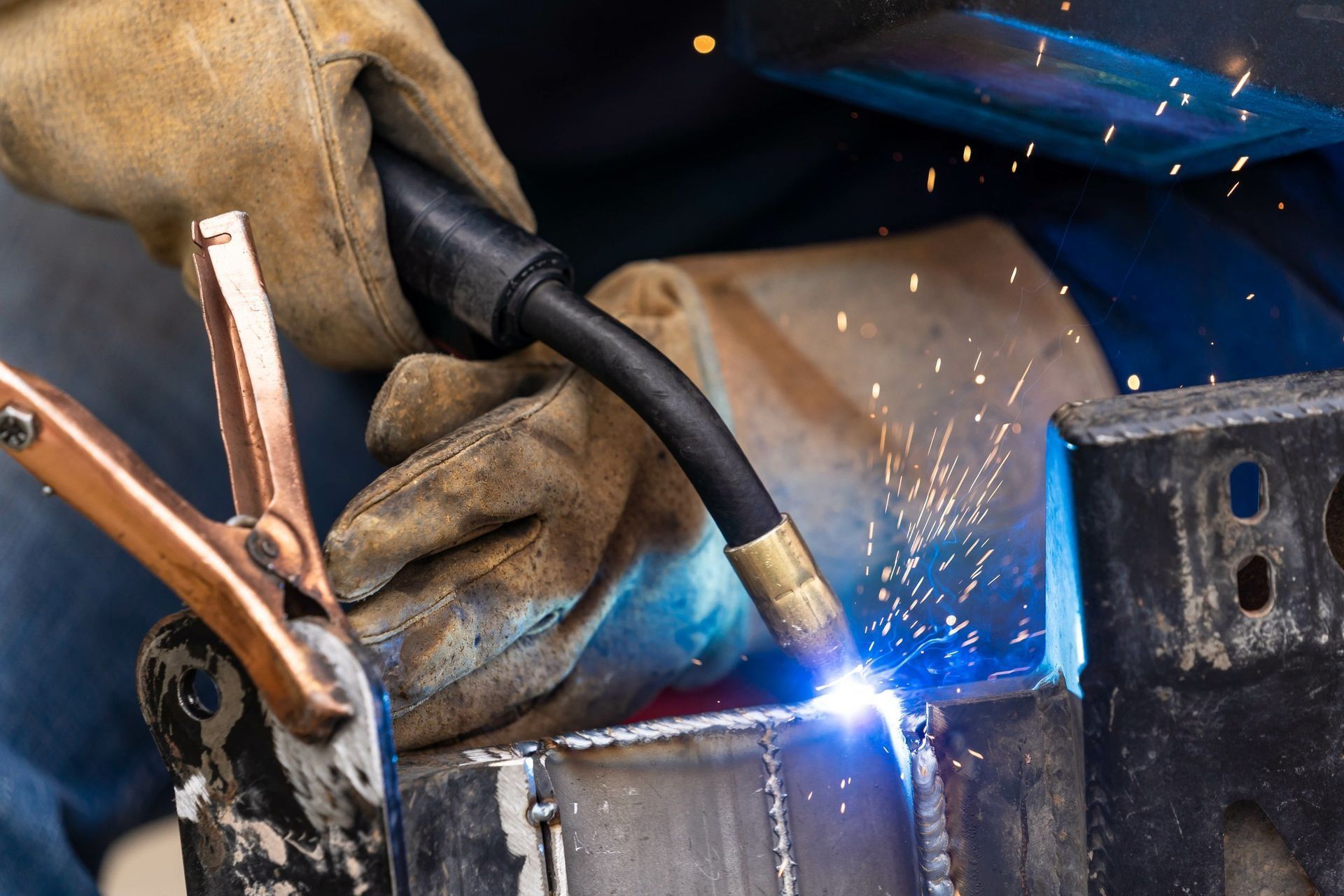 Welder in gloves using a torch on metal, with bright blue sparks flying in a workshop.