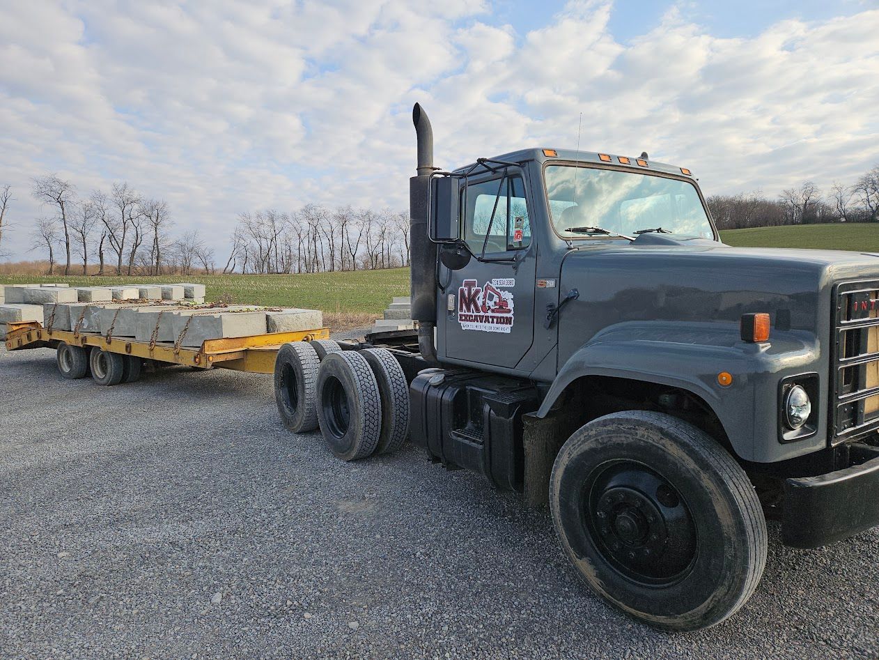 Black semi truck hauling a flatbed trailer on gravel in a rural field under cloudy sky