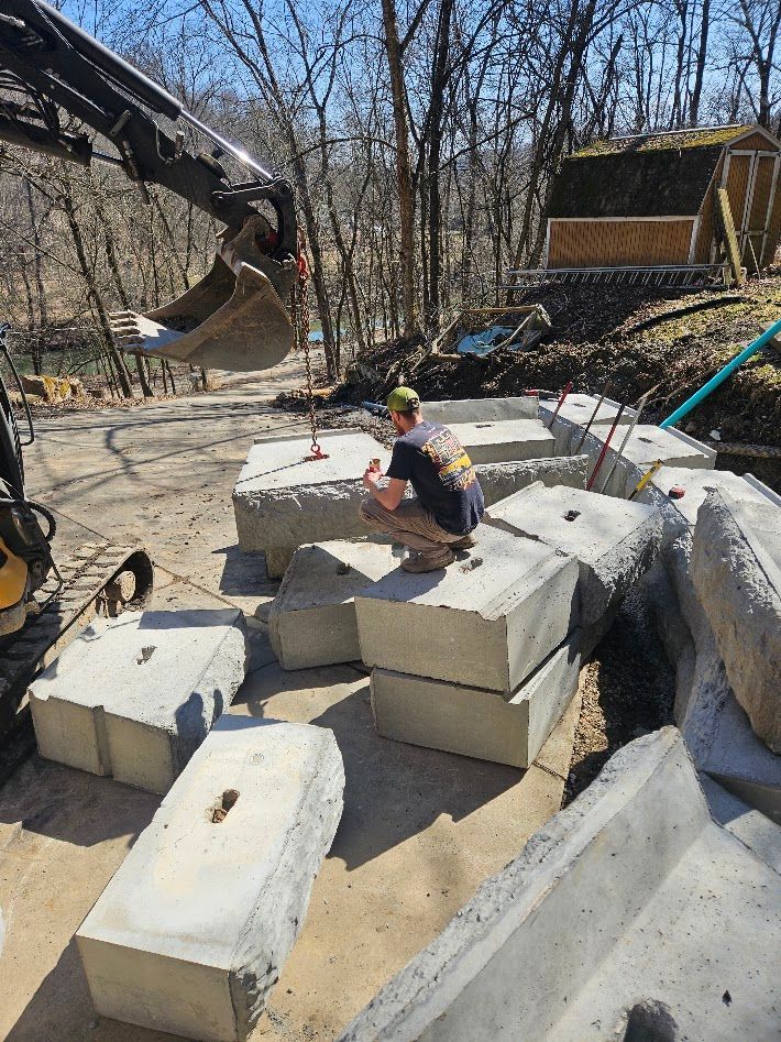 Person sitting on large concrete blocks near an excavator in a wooded outdoor worksite