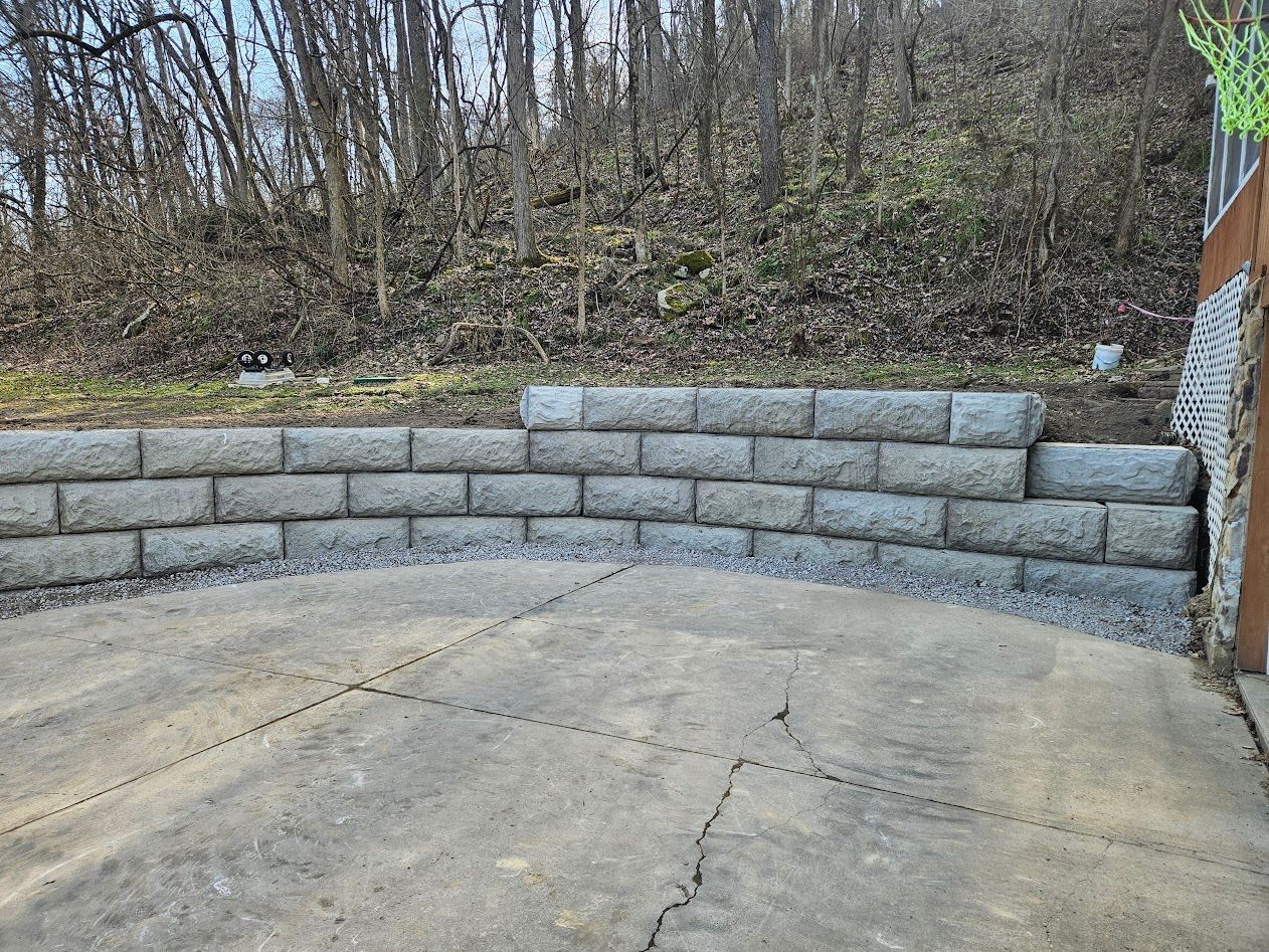 Curved gray block retaining wall around a concrete patio beside a wooded hillside