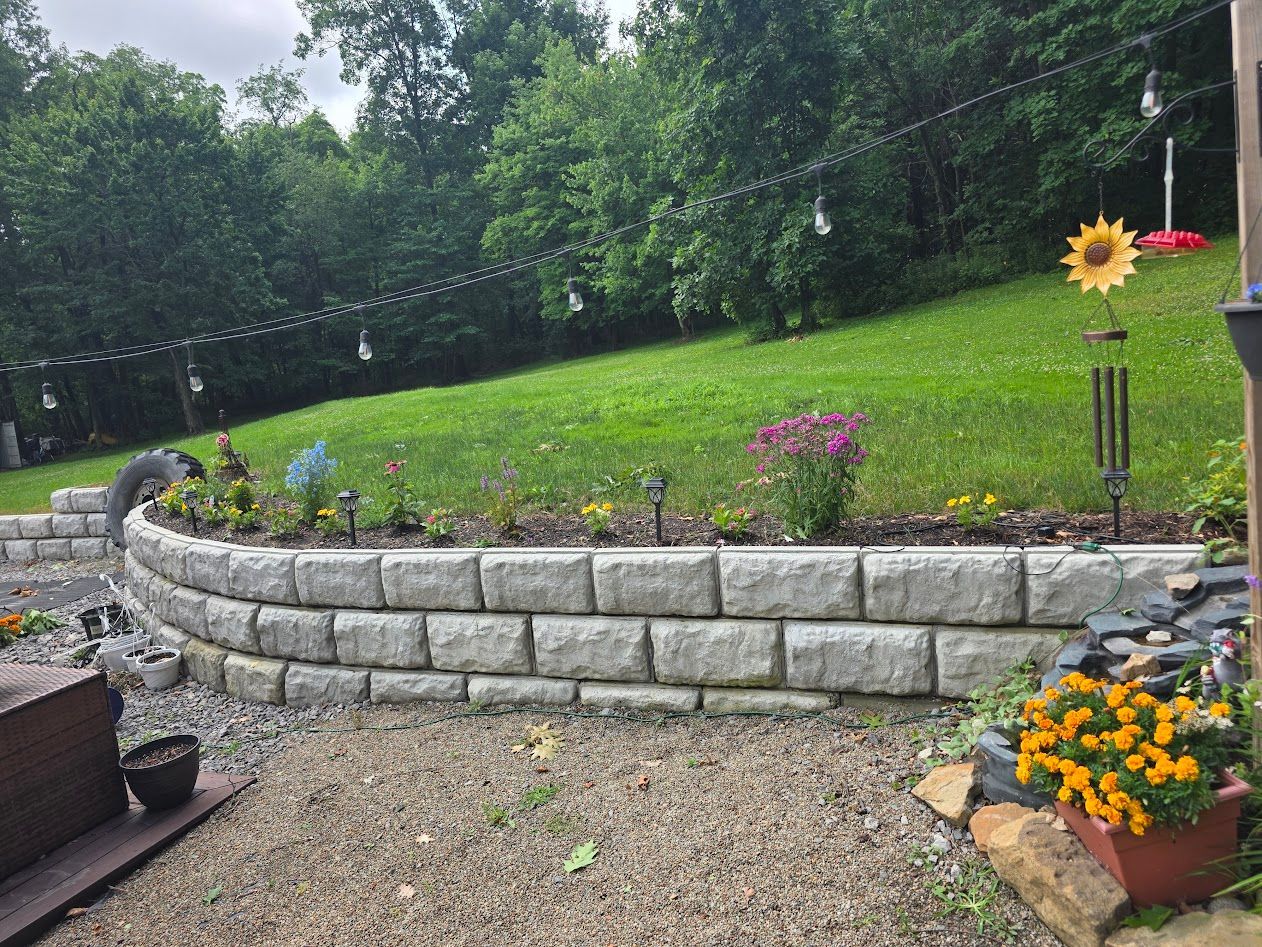 Curved stone retaining wall with flowers in a landscaped garden beside a grassy lawn and trees.