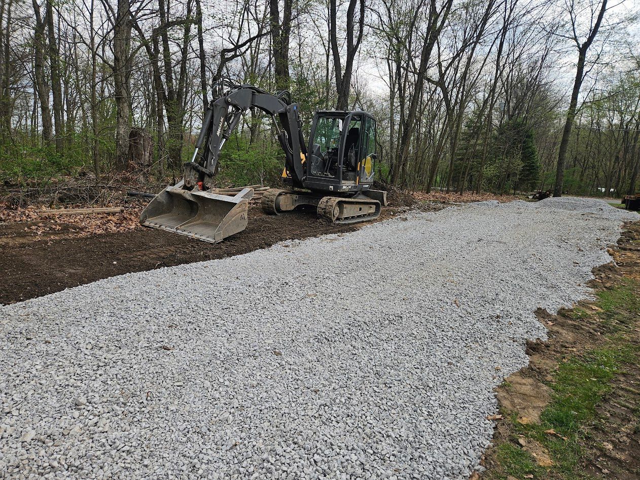 Excavator grading a gravel path in a wooded area during site work