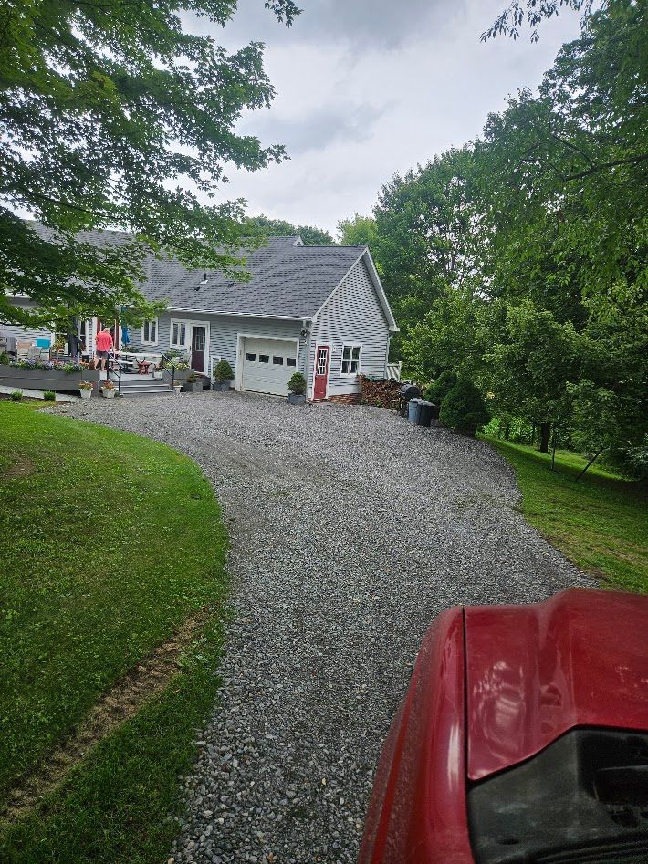 Gravel driveway leading to a white house with gray roof, surrounded by green trees on a rainy day