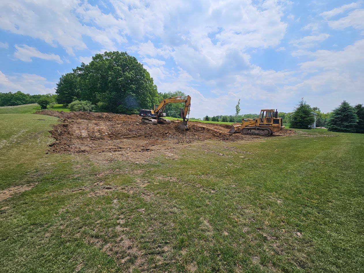 Excavator clearing a dirt mound in a grassy field under a cloudy blue sky