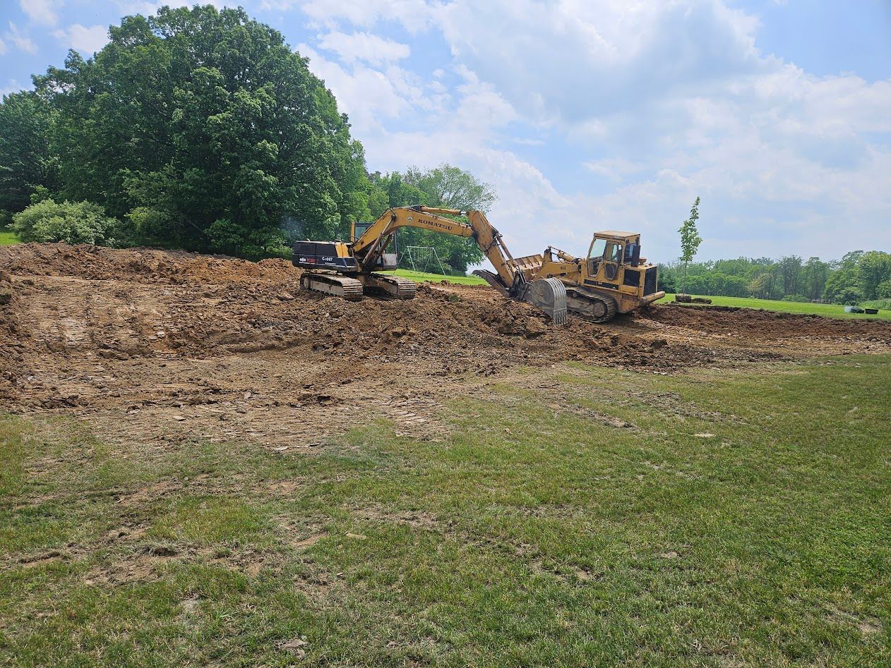Two excavators clearing a dirt mound in a grassy field with trees in the background