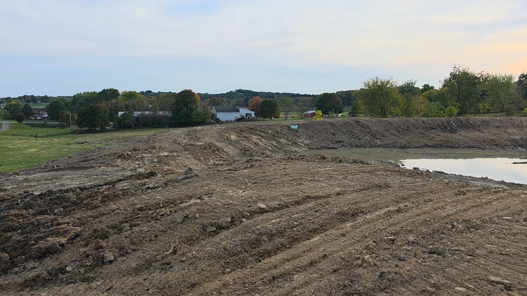 Cleared dirt lot with tire tracks beside a small pond and trees in the distance