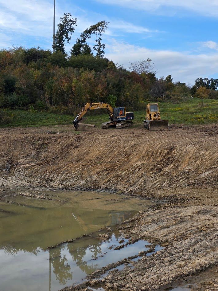 Two excavators at a muddy construction site beside a pond under a cloudy sky