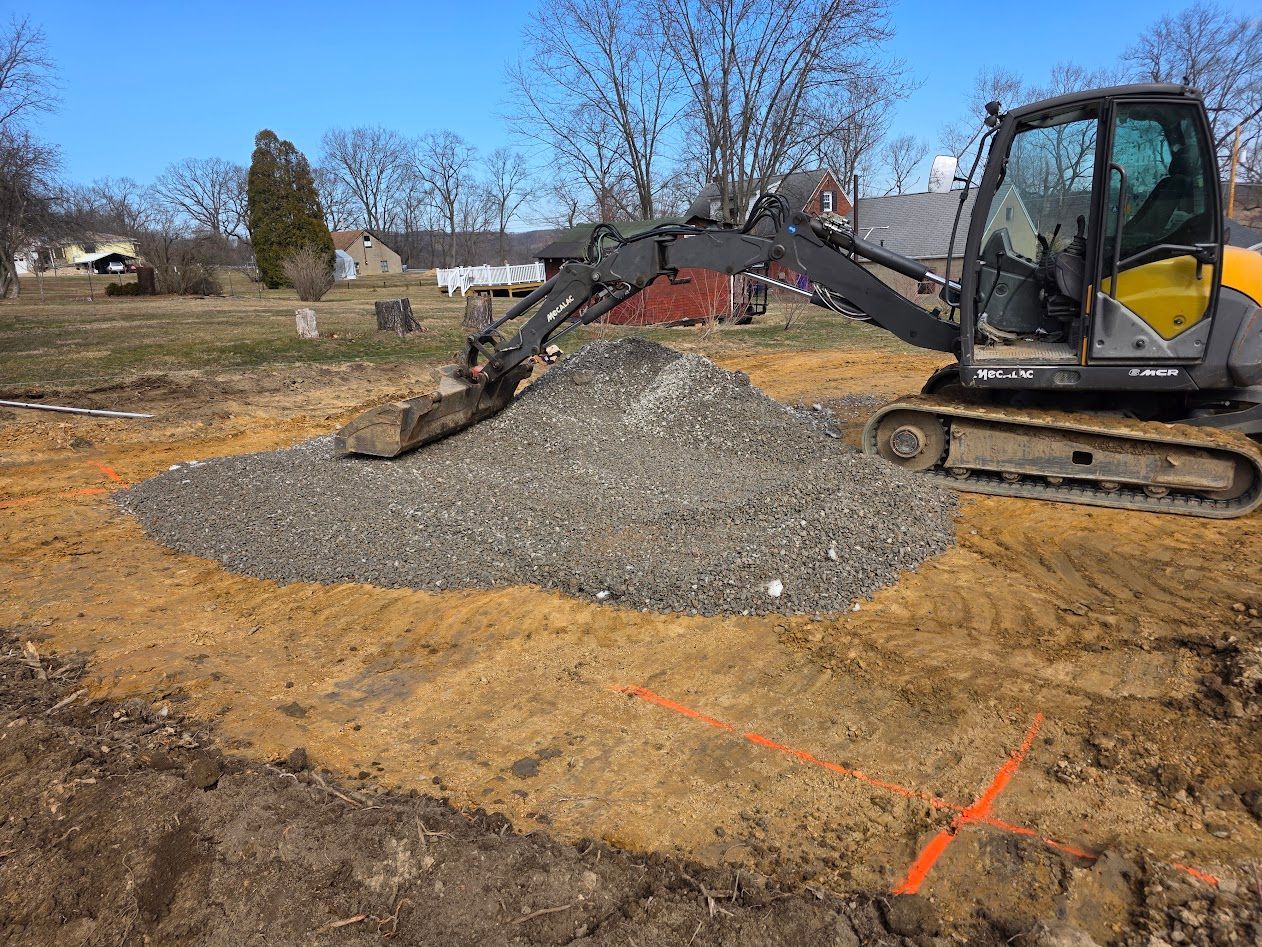 Excavator with extended arm moving gravel on a dirt lot beside a house under a clear sky