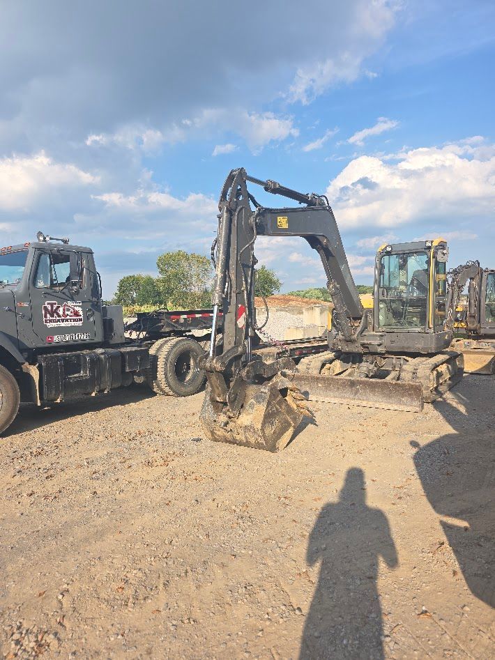 Black excavator on dirt lot beside trucks under a partly cloudy sky