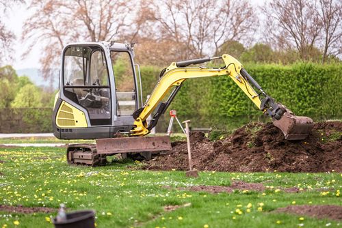 Small excavator digging soil in a grassy field beside a hedge and trees
