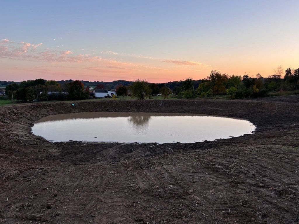 A muddy field with a pond at sunset, trees and houses in the distance.