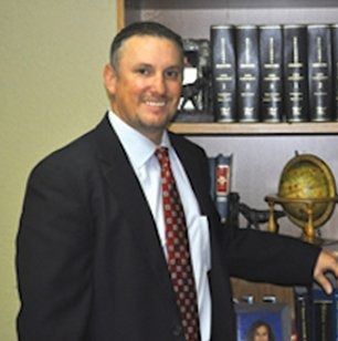 Man in suit smiles, standing by bookshelf with legal books and globe.