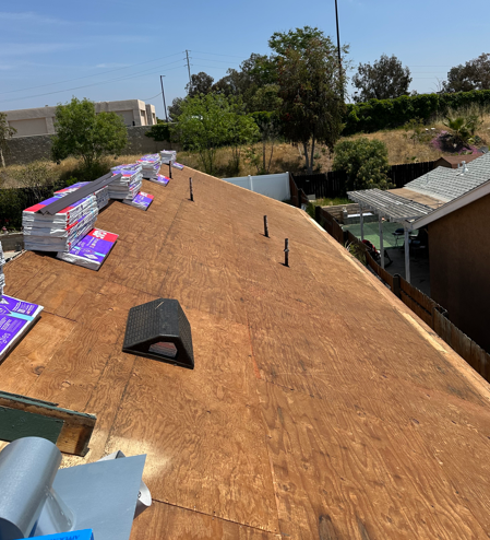 Roof under construction with plywood sheathing, stacked shingles, and a vent in bright sunlight