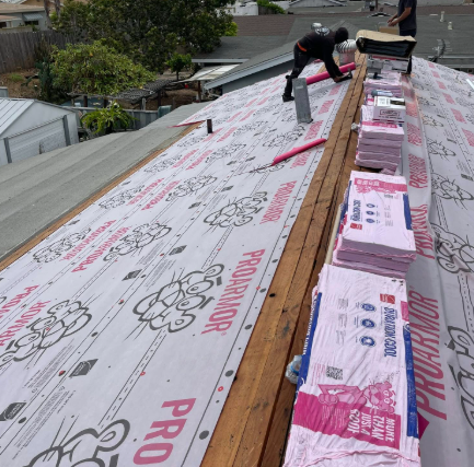 Workers installing pink underlayment on a roof, with stacked materials along the ridge.