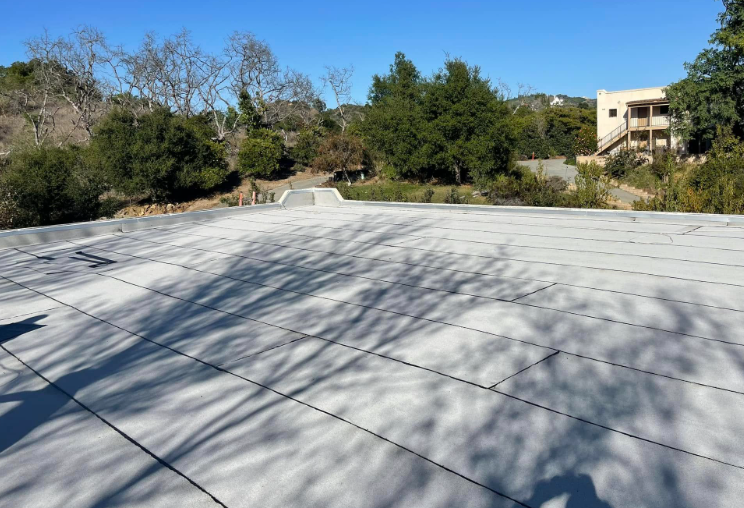 Rooftop with gray shingles and tree shadows, overlooking a sunny hillside and a building in the distance.