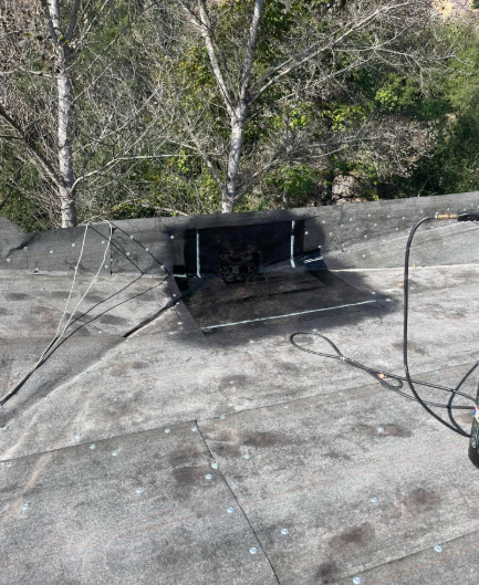 Flat rooftop with a black vent opening and cables, backed by trees and hillside.