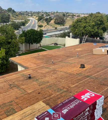Rooftop with stacked roofing materials overlooking trees and a winding road on a sunny day