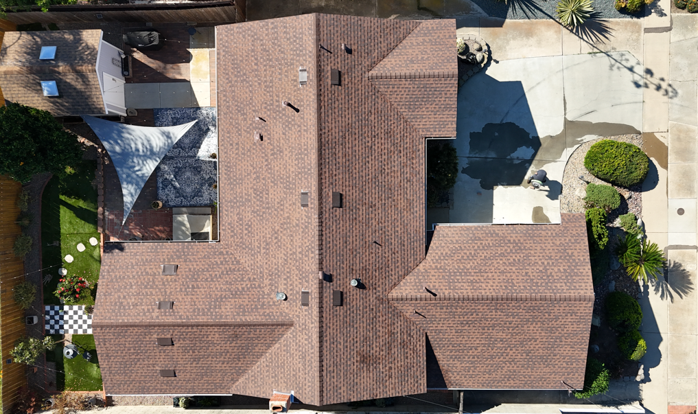 Aerial view of a house with a brown shingled roof, surrounded by a driveway, trees, and a paved patio.