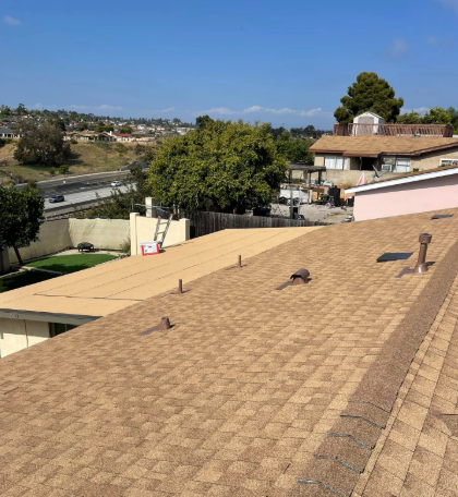 Rooftop with tan shingles overlooking trees, houses, and a blue sky on a sunny day