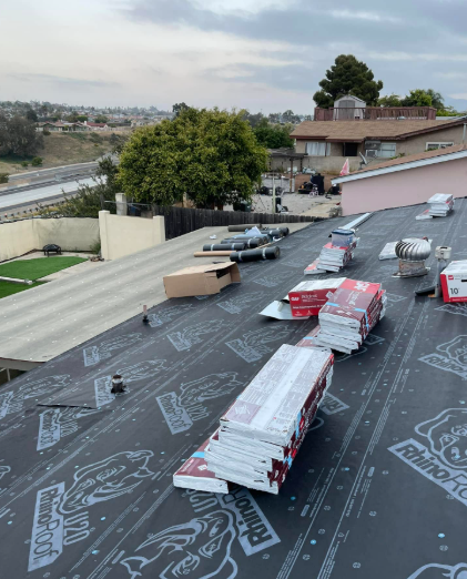 Roof under construction with black underlayment, stacks of shingles, and a parking lot below