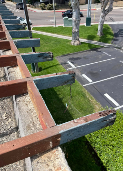 Broken wooden railing beside a grassy drainage ditch and parking lot with trees and white lane markings