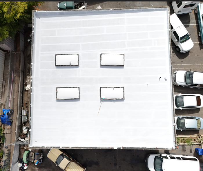 Aerial view of a white rooftop with four skylights, surrounded by parked cars and trees.