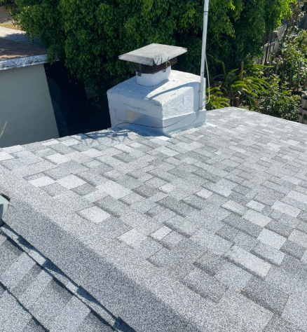 Gray shingle roof with a white chimney and vent cap beside leafy trees