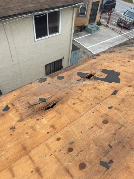 Damaged flat roof with exposed plywood and black tar patches beside a beige building