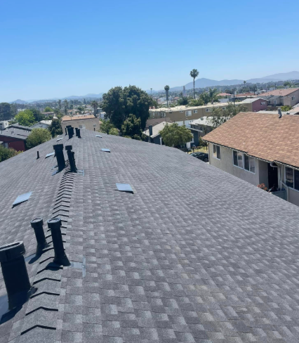Rooftop with gray shingles and vents, overlooking nearby houses and a distant city skyline under a clear blue sky
