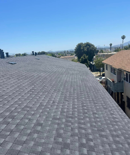 Rooftop with gray shingles beside a tan building under a clear blue sky.