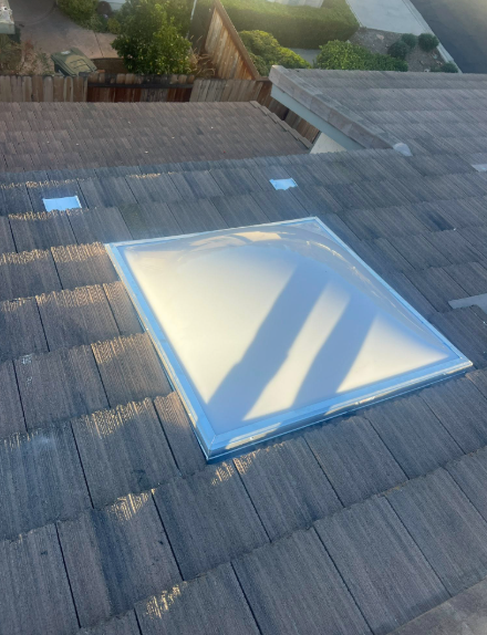Rooftop skylight on gray shingles, with sunlight casting shadows across the glass.