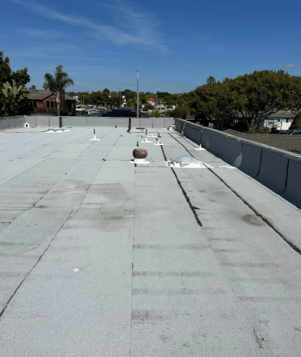 Rooftop with white coating, vents, and a clear blue sky over nearby trees and houses.