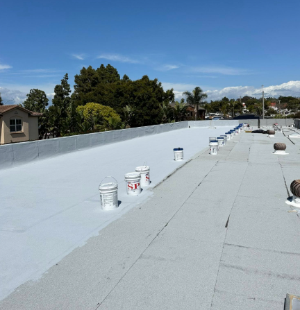 Flat rooftop with white coating and paint buckets, overlooking trees and houses under a blue sky