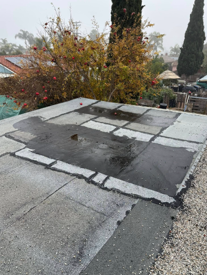 Flat rooftop with patched gray tar paper and a wet surface, overlooking trees and a pool in the background