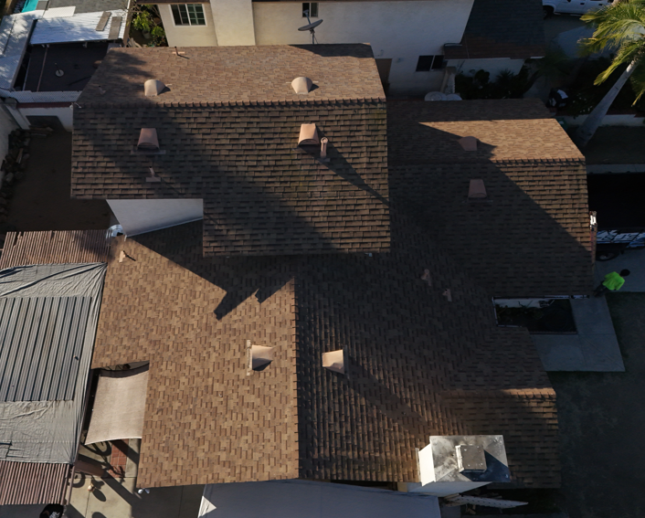 Aerial view of a brown shingled house roof with vents and skylights in a residential neighborhood